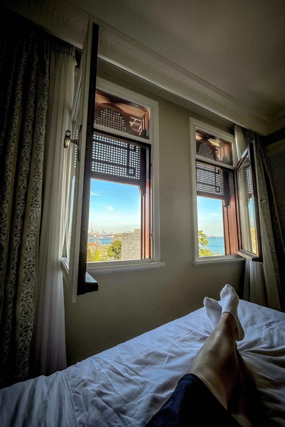 A relaxed view from a Istanbul hotel bed with feet stretched out, looking through open wooden windows at a bright blue sky and the sea in the distance.