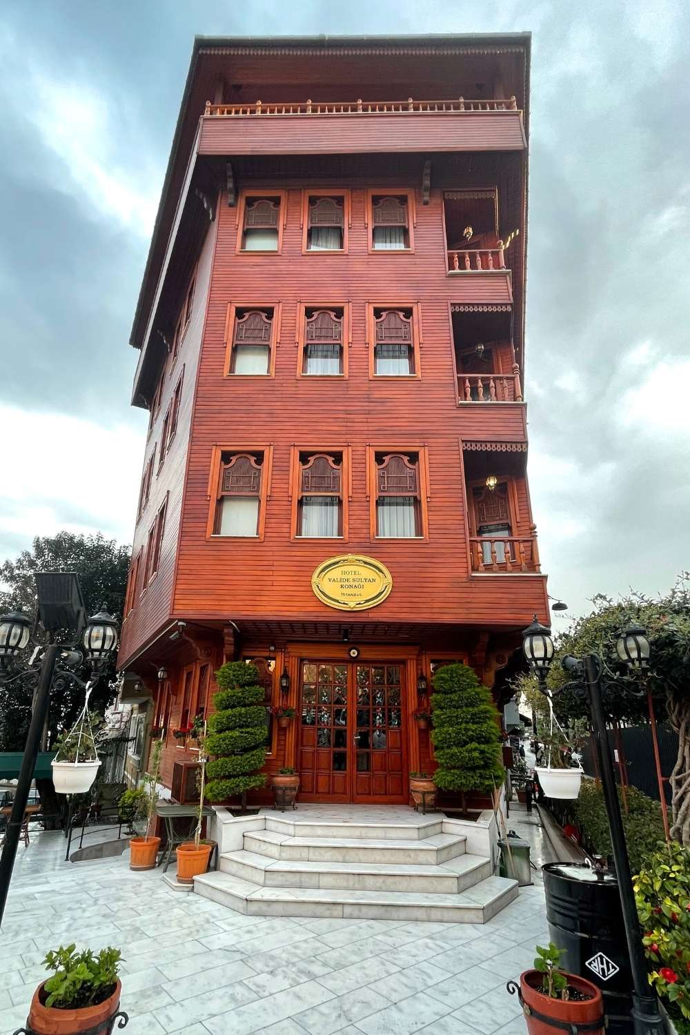 A traditional Ottoman-style wooden hotel, "Valide Sultan Konagi," stands tall with a deep red facade, ornate shutters, and a grand staircase entrance surrounded by greenery and lanterns under an overcast sky.