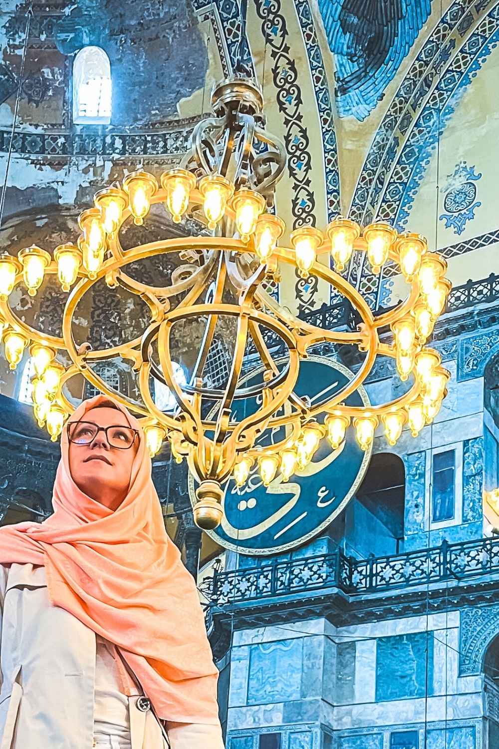 Kate in a peach-colored hijab and glasses, gazes upward inside the Hagia Sophia, standing beneath an ornate golden chandelier, with Islamic calligraphy and intricate Byzantine architecture in the background.