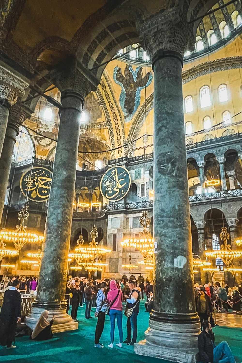 A vibrant scene inside the Hagia Sophia in Istanbul, showcasing towering marble columns, golden chandeliers, Islamic calligraphy, and a Byzantine angel mosaic on the dome, with visitors exploring the historic site.