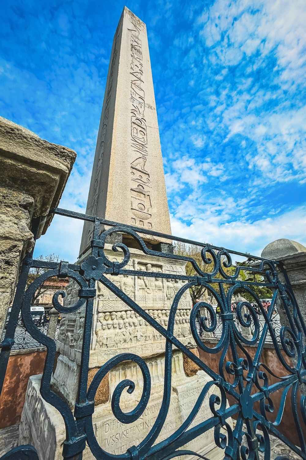 The Obelisk of Theodosius in Istanbul, an ancient Egyptian monument with intricate carvings, stands tall against a bright blue sky, framed by an ornate wrought iron fence in the foreground.