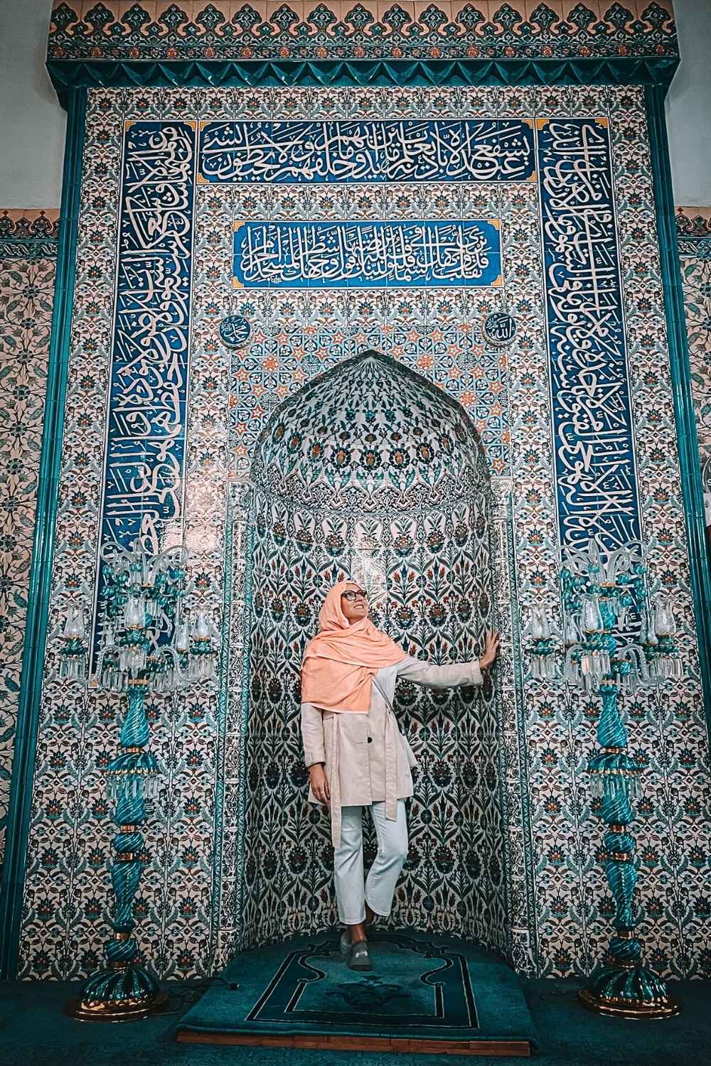 Kate in a peach hijab and beige coat stands gracefully in front of an ornate mihrab adorned with intricate blue and white Iznik tiles and Arabic calligraphy, inside a historic mosque in Istanbul.