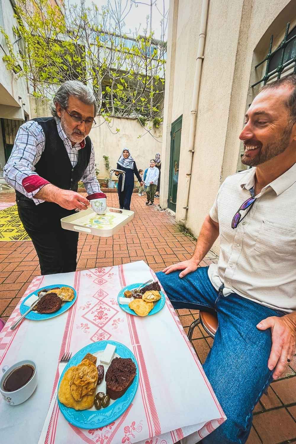 Kate's husband sits at an outdoor table in a courtyard in Istanbul, as an elderly local man in a vest and checkered shirt serves traditional Turkish tea, with plates of pastries, cheese, and olives in front of him and a family in the background.