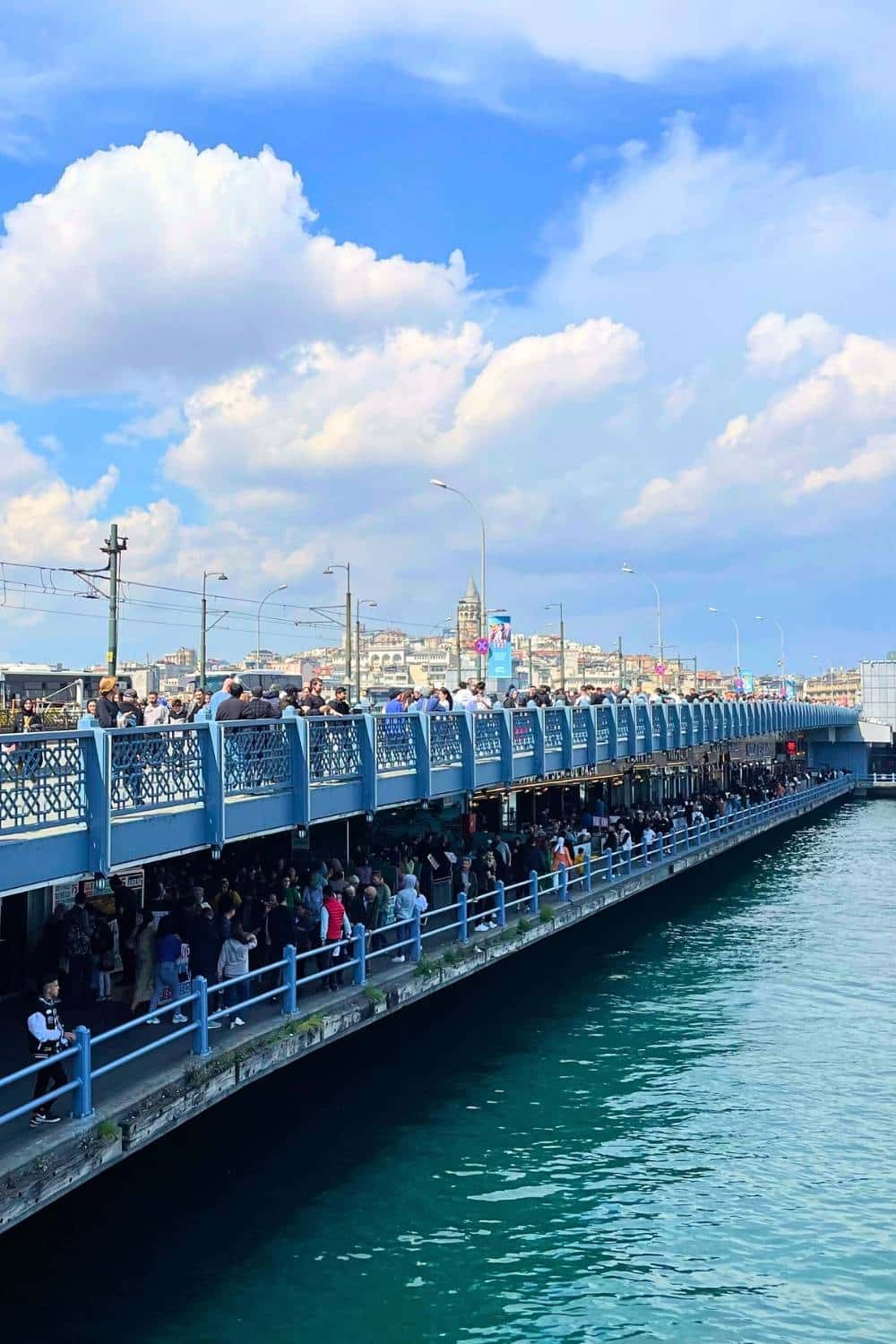 The bustling Galata Bridge in Istanbul, filled with pedestrians and fishermen on the upper level, while crowds explore the waterfront restaurants and shops below, with the historic Galata Tower visible in the background under a bright blue sky.