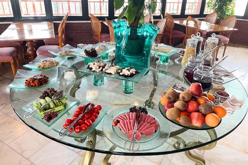 A beautifully arranged Turkish breakfast spread on a round glass table, featuring fresh fruits, vegetables, cheeses, cold cuts, olives, jams, and breads, set in a sunlit dining area with wooden chairs and large windows.