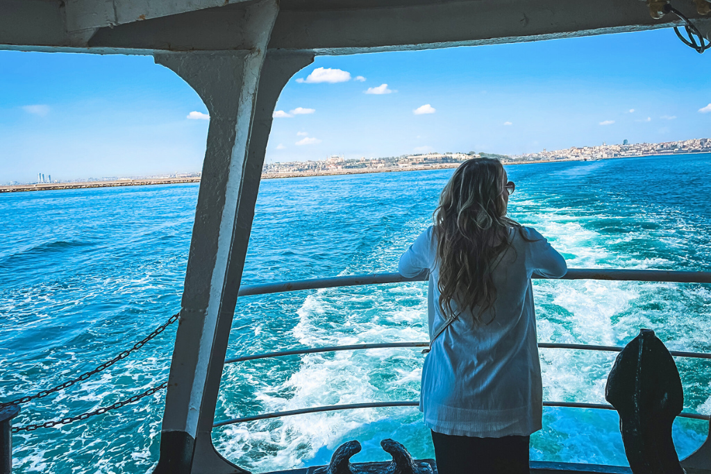 Kate with long wavy blonde hair leans against the railing of a ferry, gazing at the deep blue waters of the Bosphorus Strait and distant shoreline under a bright sky.