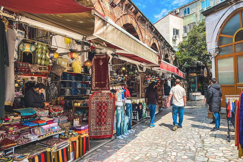 A bustling outdoor market in Istanbul with colorful textiles, decorative lamps, and traditional goods on display, as shoppers stroll along the mosaic-tiled walkway under shaded awnings.