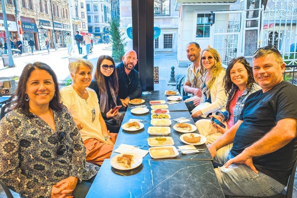 A group of smiling people on a food tour, gathers around a table at an outdoor café in Istanbul, enjoying a traditional Turkish breakfast, with a lively street scene in the background.