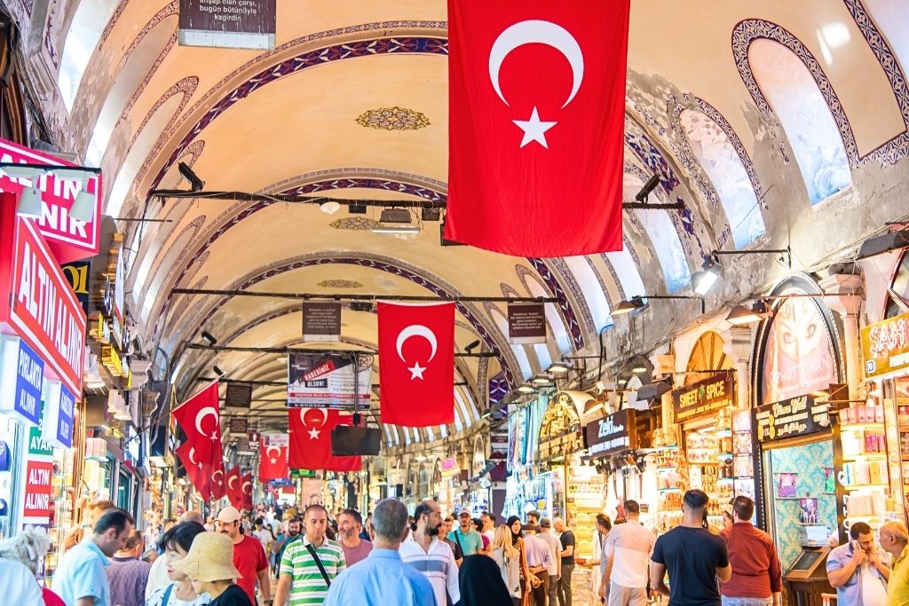 The bustling Grand Bazaar in Istanbul, with arched ceilings adorned with Turkish flags, vibrant shop signs, and crowds of locals and tourists exploring the historic marketplace.