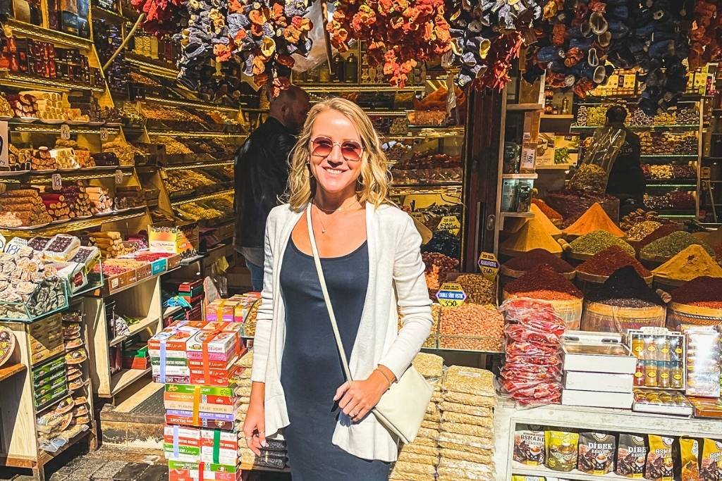 Kate in sunglasses and a black dress stands in front of a vibrant spice and sweets shop in Istanbul, surrounded by shelves of Turkish delights, dried fruits, nuts, and colorful spice pyramids.