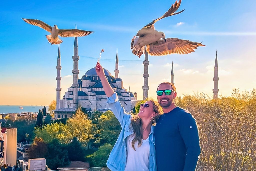 Kate in a denim jacket raises food for seagulls as they soar over a rooftop with the Blue Mosque in the background. She stands beside her husband in sunglasses, both smiling.