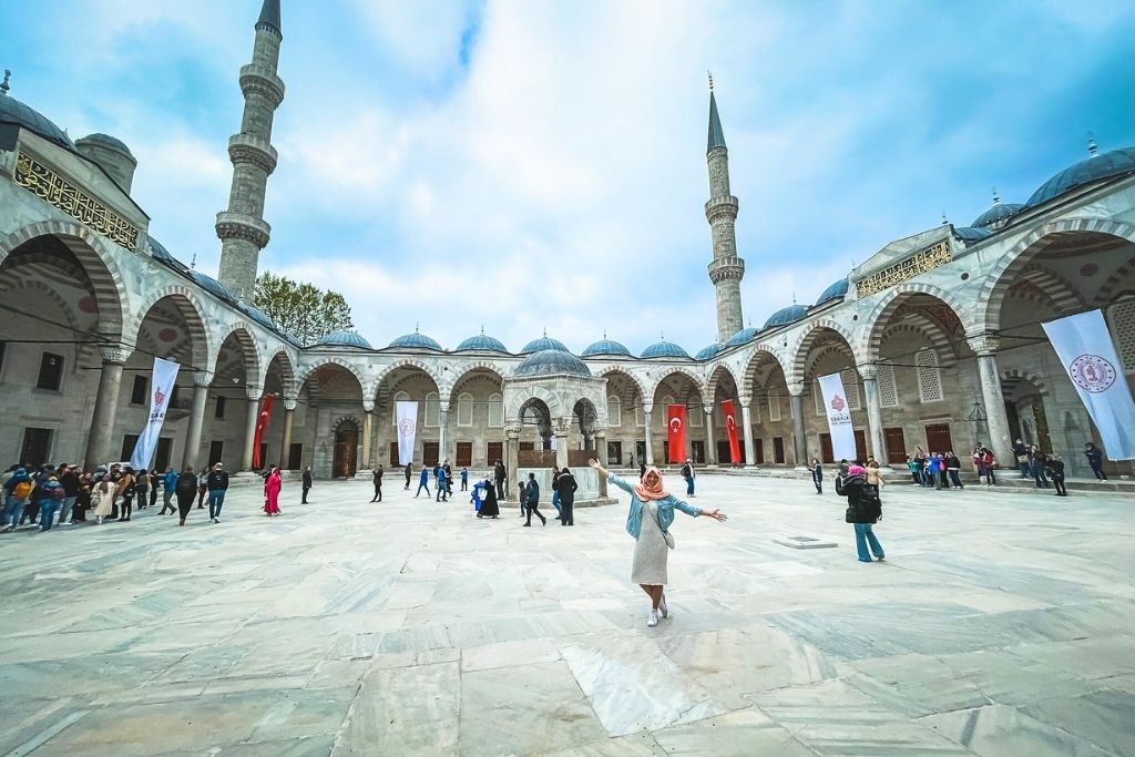 A grand courtyard of the Blue Mosque, surrounded by arched colonnades and towering minarets, with a Kate in the center joyfully extending her arms as visitors explore the site.