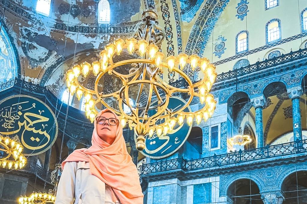 Kate, in a peach-colored hijab and glasses, stands inside the Hagia Sophia in Istanbul, illuminated by grand golden chandeliers with intricate Islamic calligraphy and ornate domes in the background.