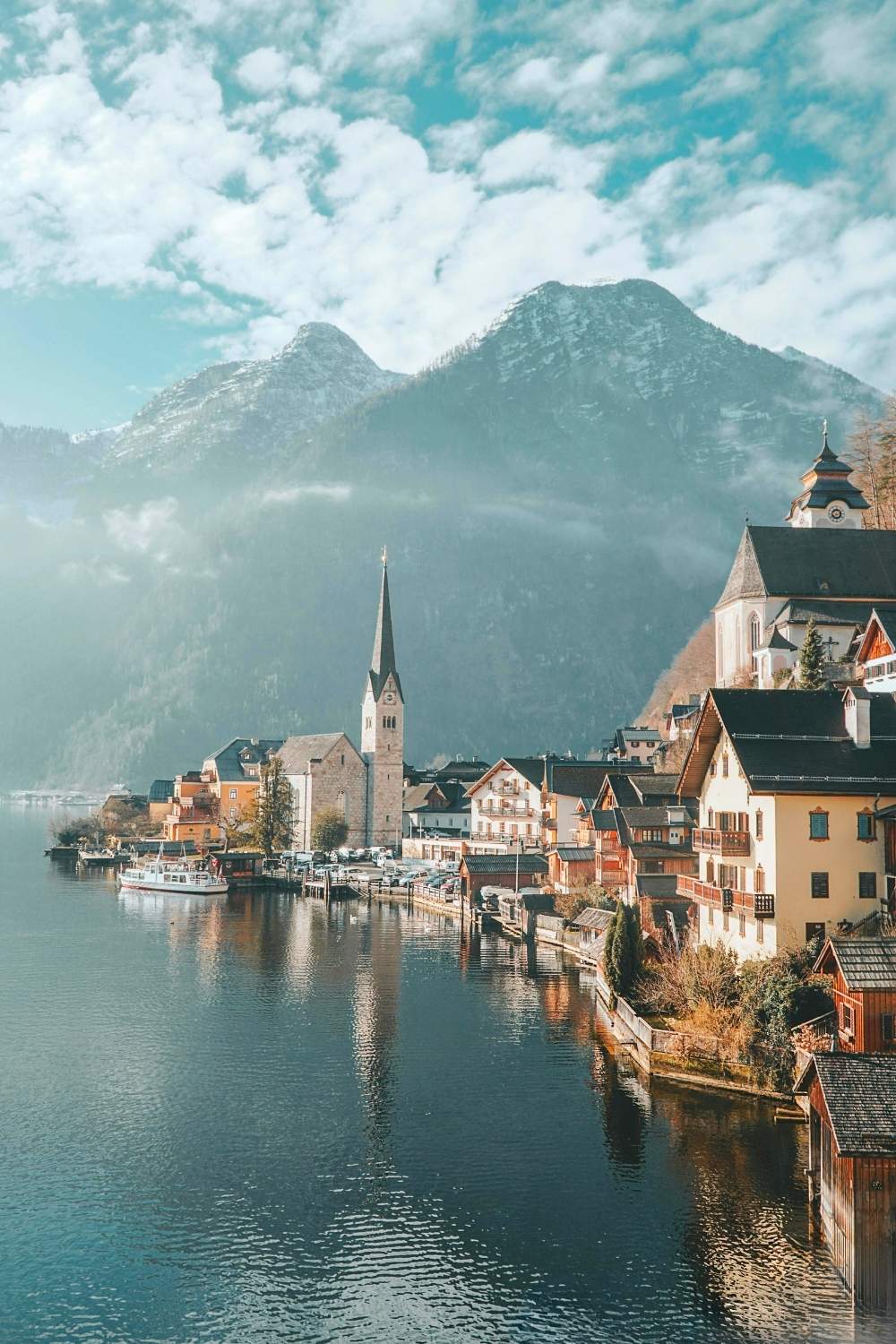 This picturesque image captures the charming lakeside village of Hallstatt, Austria, with its iconic church spire, traditional alpine houses, and serene waters reflecting the scene. Towering snow-capped mountains frame the background under a vibrant, partly cloudy sky, creating a dreamy and tranquil atmosphere.