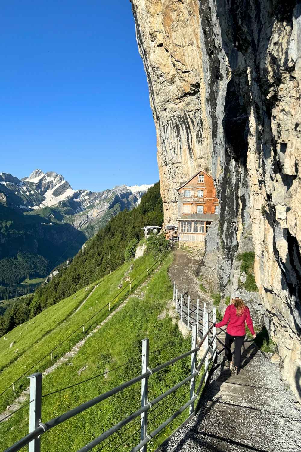 This image features the iconic Aescher Mountain Guesthouse nestled against a sheer cliff in Appenzell, Switzerland. Kate, in a bright pink jacket, walks along a narrow trail leading to the guesthouse, with lush green hills and snow-capped mountains in the background under a clear blue sky.