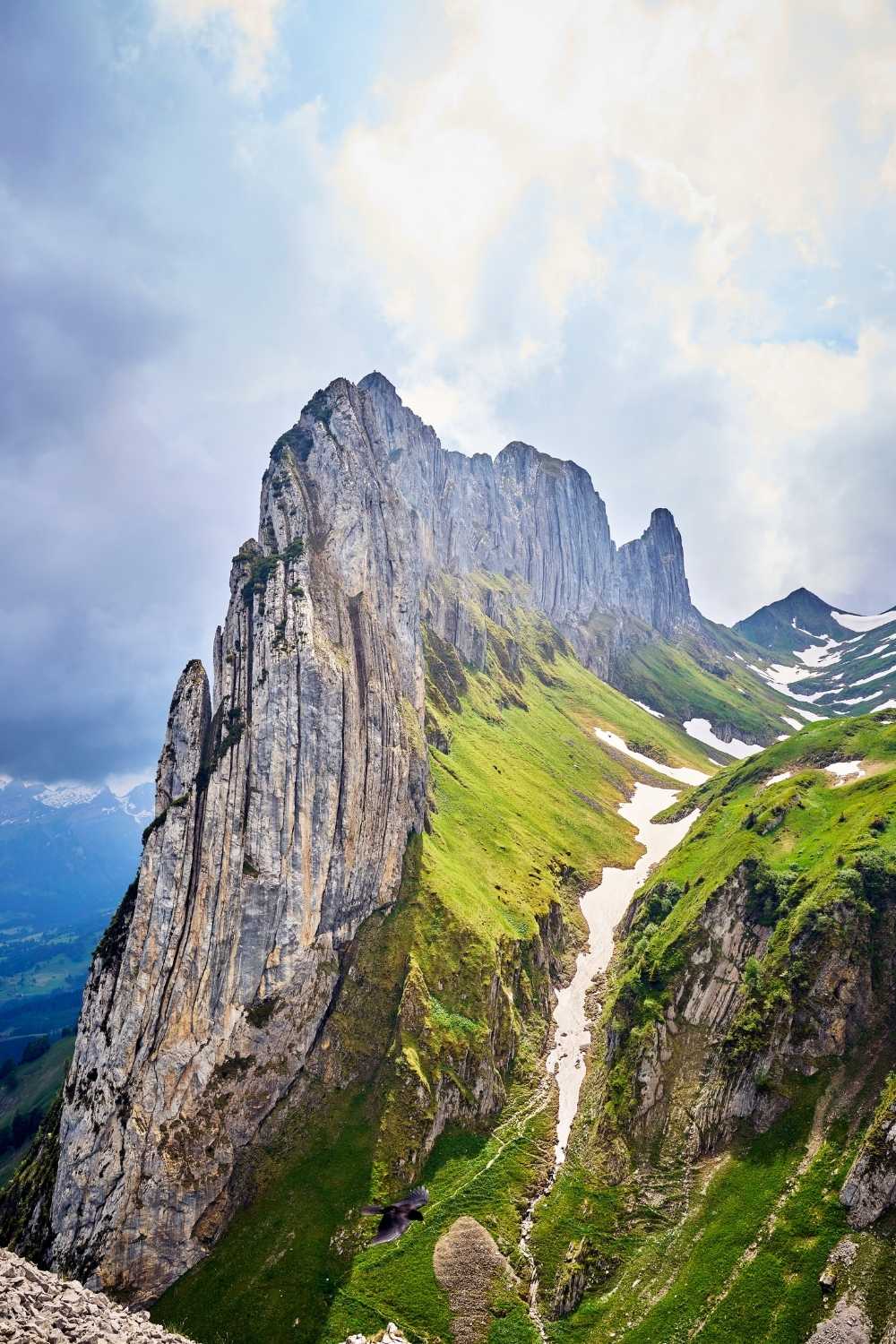 This stunning image showcases the dramatic Aescher Ridge in Switzerland, with its towering, jagged cliffs and lush green slopes. Snow patches are scattered along the ridges, adding contrast to the vibrant landscape under a partly cloudy sky. The majestic natural formation highlights the rugged beauty of the Swiss Alps.