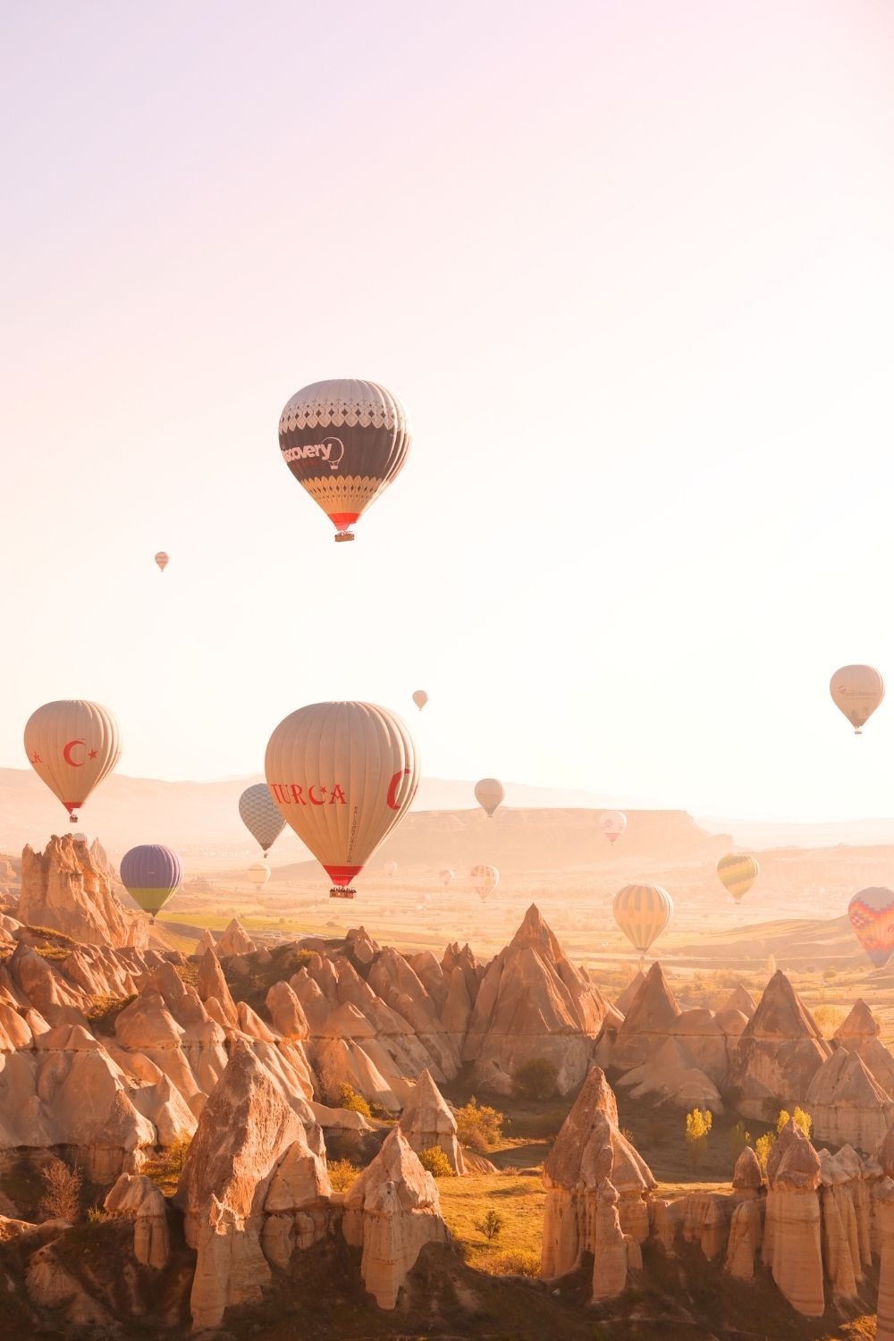 This image showcases the enchanting sight of hot air balloons floating above the unique rock formations of Cappadocia, Turkey, during a golden sunrise. The soft pastel hues of the sky and the warm light illuminating the fairy chimneys create a magical and serene atmosphere. The scene captures the timeless beauty and allure of Cappadocia's iconic landscape.