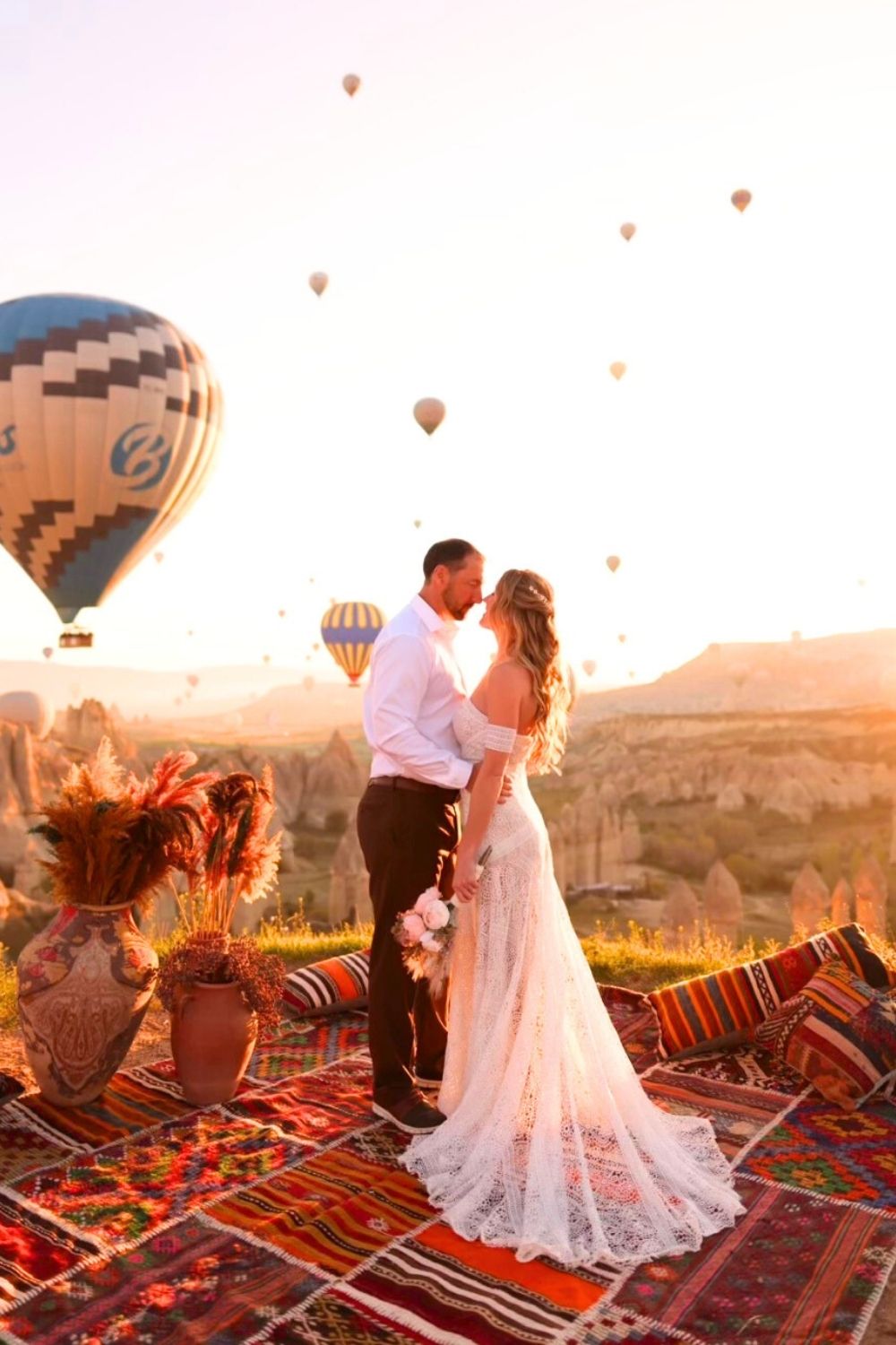 This image captures a romantic moment in Cappadocia, Turkey, where a couple stands on a colorful patterned rug, surrounded by traditional vases filled with dried flowers. They embrace as hot air balloons float above the unique rock formations in the warm glow of sunrise. The bride's lace gown and bouquet add elegance to the dreamy and picturesque setting.