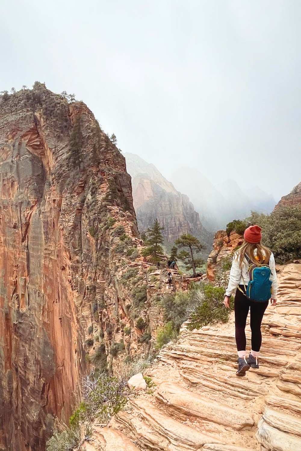 This image captures Kate on the thrilling Angels Landing trail in Zion National Park, Utah, surrounded by towering red rock cliffs and rugged terrain. The pathway is narrow and steep, offering breathtaking views of the canyon below shrouded in mist. The scene highlights the adventurous and dramatic nature of this iconic hike.