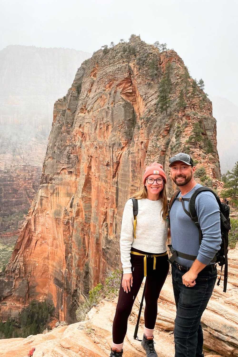 This image features a smiling Kate and her husband standing on the Angels Landing trail in Zion National Park, Utah, with the iconic red rock cliffs towering behind them. Both are equipped with backpacks and hiking gear, highlighting their adventurous spirit. The misty backdrop adds depth and drama to this breathtaking location.