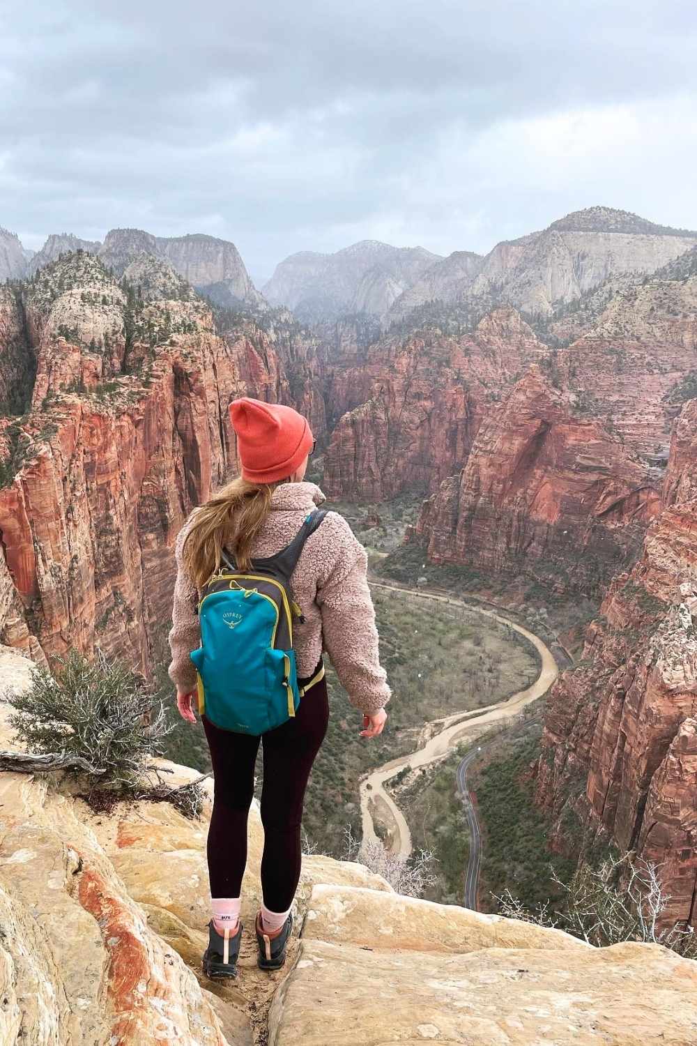 This image depicts Kate standing at the edge of a cliff on Angels Landing in Zion National Park, Utah, overlooking the breathtaking red rock canyon and the winding river below. Wearing a cozy jacket, orange beanie, and a teal backpack, the hiker gazes at the expansive and dramatic landscape under a cloudy sky. 