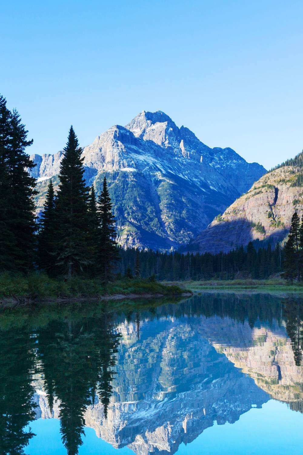 This image captures a serene mountain scene in Glacier National Park, featuring a towering, rugged peak with snow-dusted slopes reflected perfectly in a calm lake below. Tall evergreen trees frame the foreground, enhancing the natural beauty of the landscape. The clear blue sky and pristine water create a tranquil and majestic atmosphere.