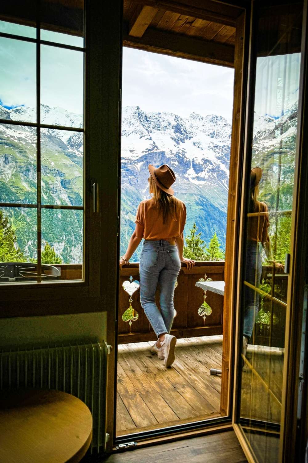 This image captures a serene moment in Mürren, Switzerland, where Kate stands on a wooden balcony gazing at the breathtaking snow-capped mountains of the Swiss Alps. Framed by a rustic door and window, the scene combines cozy indoor warmth with the grandeur of nature outside.