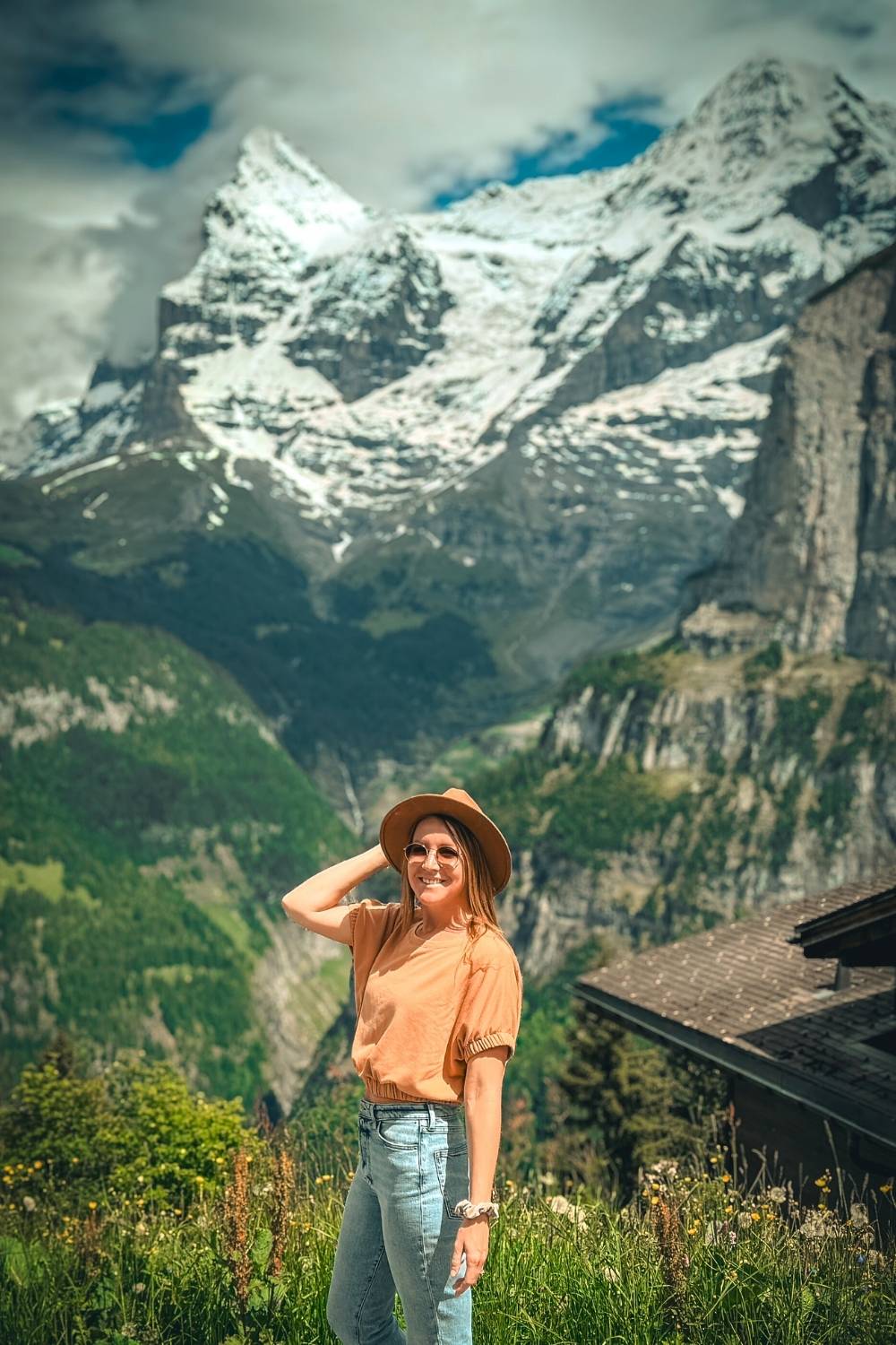 This image features Kate smiling in Mürren, Switzerland, standing against a backdrop of snow-capped peaks and lush green alpine valleys. She wears a stylish hat, sunglasses and an orange shirt with blue jeans.