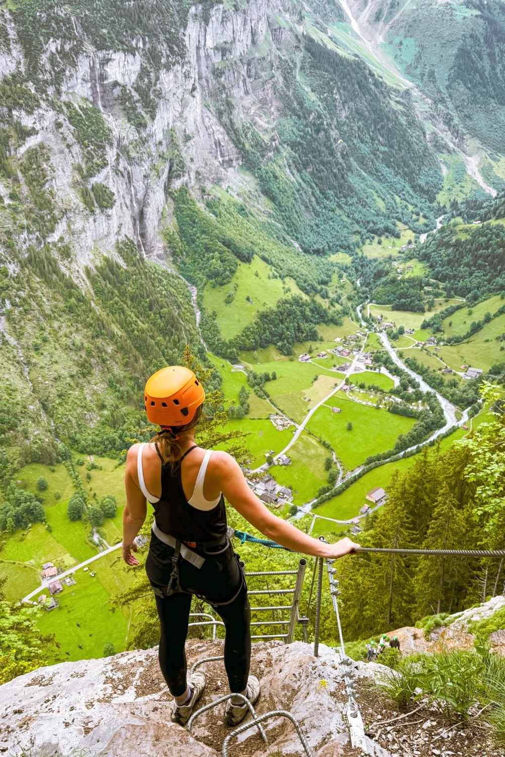 This image shows Kate equipped with a helmet and harness on the via Ferrata route in Mürren, Switzerland. Kate stands on a ladder-like structure, overlooking a breathtaking valley filled with lush green pastures, winding roads, and scattered alpine homes. Towering cliffs and dense forests surround the dramatic landscape