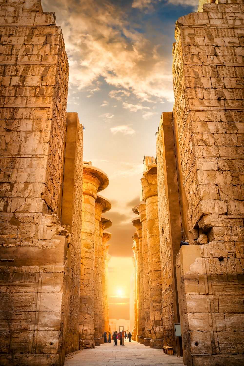 This image showcases the grand columns and ancient walls of Karnak Temple in Egypt, illuminated by the golden hues of a setting sun. Hieroglyphs intricately carved into the stone detail the rich history of this iconic archaeological site. Visitors walking through the temple emphasize its immense scale and timeless beauty.






