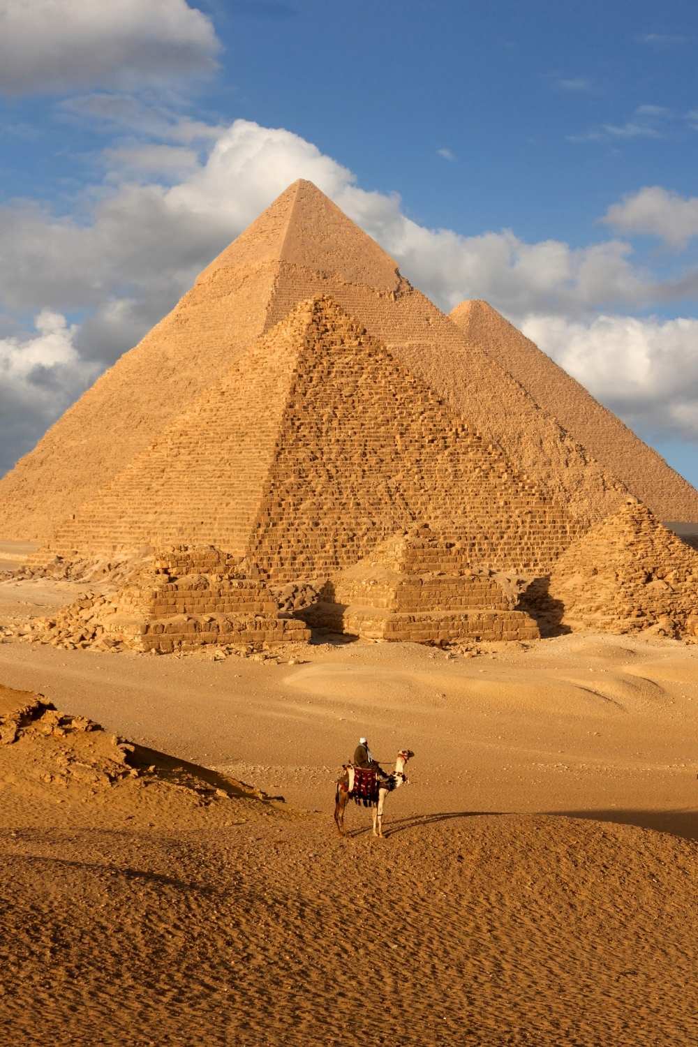 This image features the iconic Pyramids of Giza in Egypt, standing majestically under a bright blue sky with scattered clouds. A lone figure on a camel, dressed in traditional attire, adds scale and a sense of timelessness to the vast desert landscape. The golden sunlight enhances the warm hues of the pyramids and surrounding sands, creating a striking and classic view of this ancient wonder.