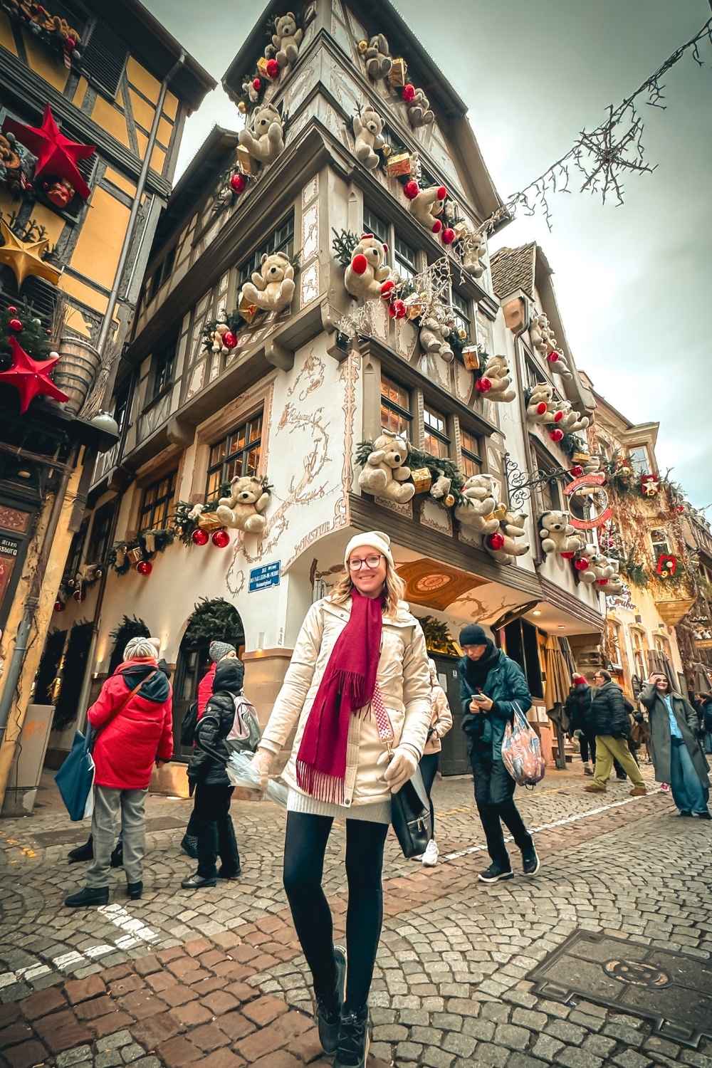 This image captures a festive street in Strasbourg, France, with a smiling Kate dressed warmly in a cream coat, red scarf, and hat. Behind her, a charming half-timbered building is adorned with oversized teddy bears, ornaments, and holiday decorations, embodying the magic of Strasbourg's famous Christmas market.