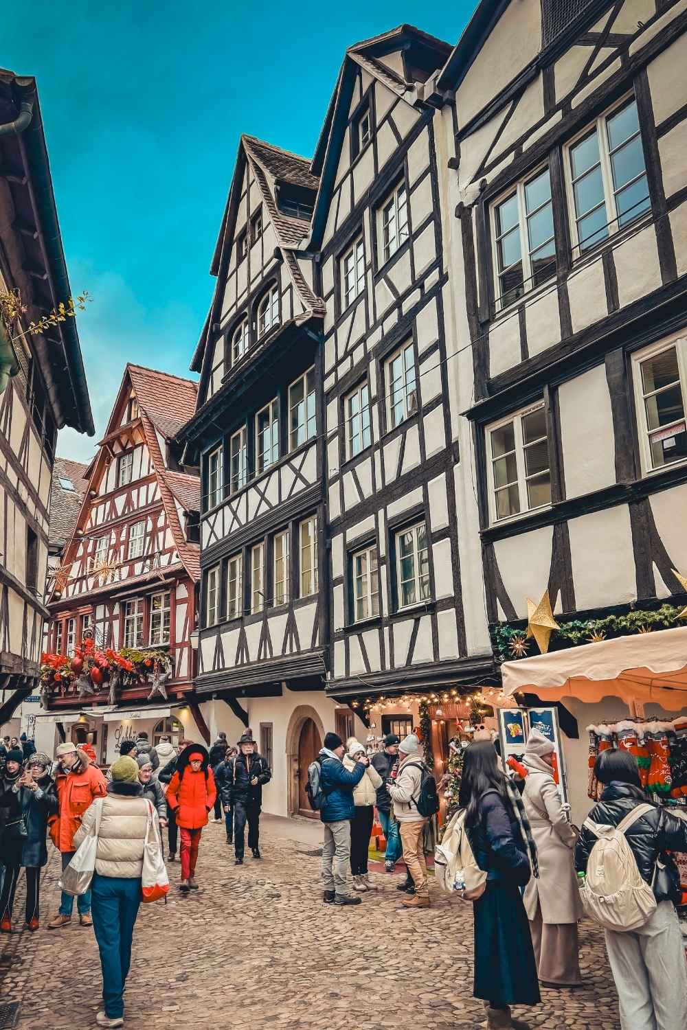 This image captures the charming streets of Petite France in Strasbourg, showcasing traditional half-timbered houses with intricate designs and sloped roofs. The cobblestone streets are lively with people exploring holiday markets adorned with festive decorations and lights. 