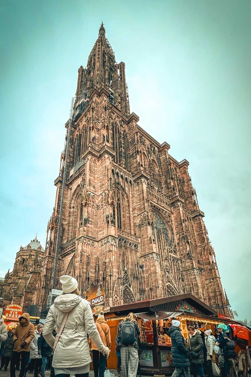 This image captures the towering Strasbourg Cathedral during the Christmas season, with its intricate Gothic architecture reaching into the sky. Festive market stalls below glow with holiday lights, and people wrapped in winter attire browse the offerings. The scene combines the grandeur of the historic cathedral with the warmth and charm of Strasbourg's famous Christmas market.