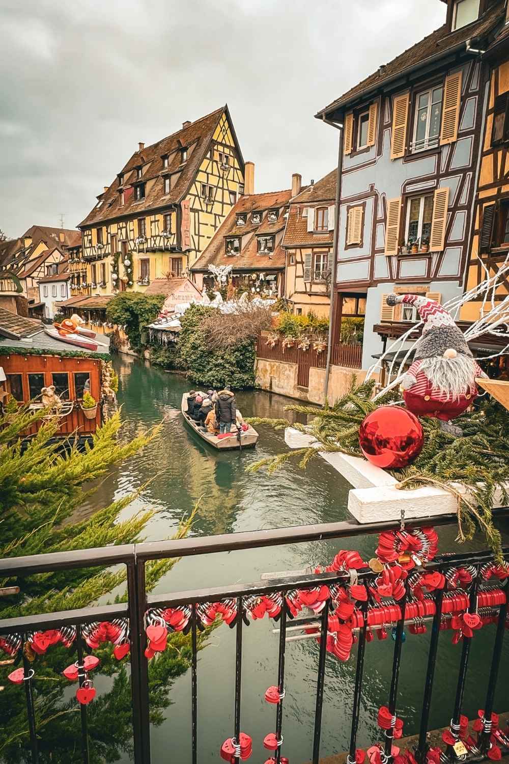 This image captures the enchanting canals of Colmar, France, during the festive season. Half-timbered houses line the water, decorated with garlands and ornaments, while a small boat glides through the canal carrying warmly dressed passengers. The foreground features a bridge adorned with red heart-shaped locks and Christmas decorations, adding to the charm and holiday spirit of this picturesque town.