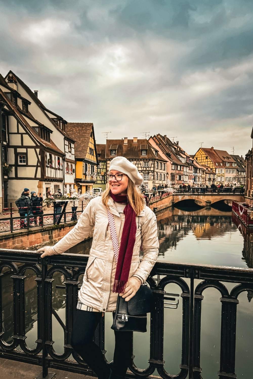 This image features a smiling Kate standing on a bridge in Colmar, France, with the town’s iconic half-timbered houses lining the canal in the background. Dressed in a cream coat, red scarf, and beret, she embodies the cozy charm of the festive season. 