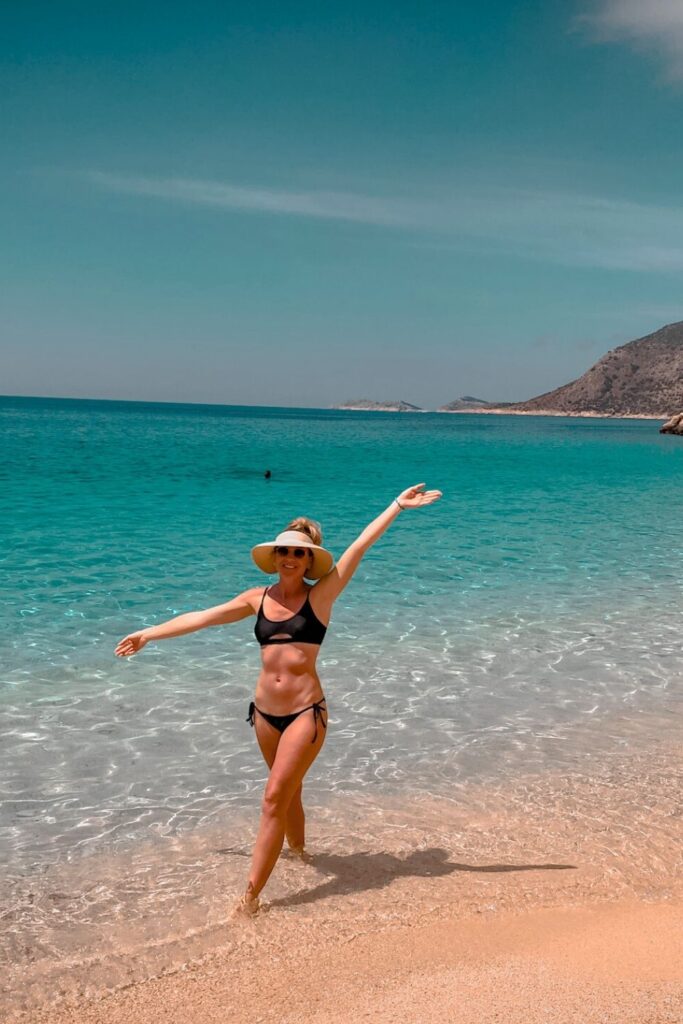 This image captures Kate on Kaputaş Beach in Turkey, featuring a woman in a black bikini and sun hat standing on the golden sand with crystal-clear turquoise water behind her. The serene beach scene is framed by a distant rugged coastline under a bright, cloudless sky. The vibrant colors and relaxed atmosphere highlight the beauty of this idyllic seaside destination.