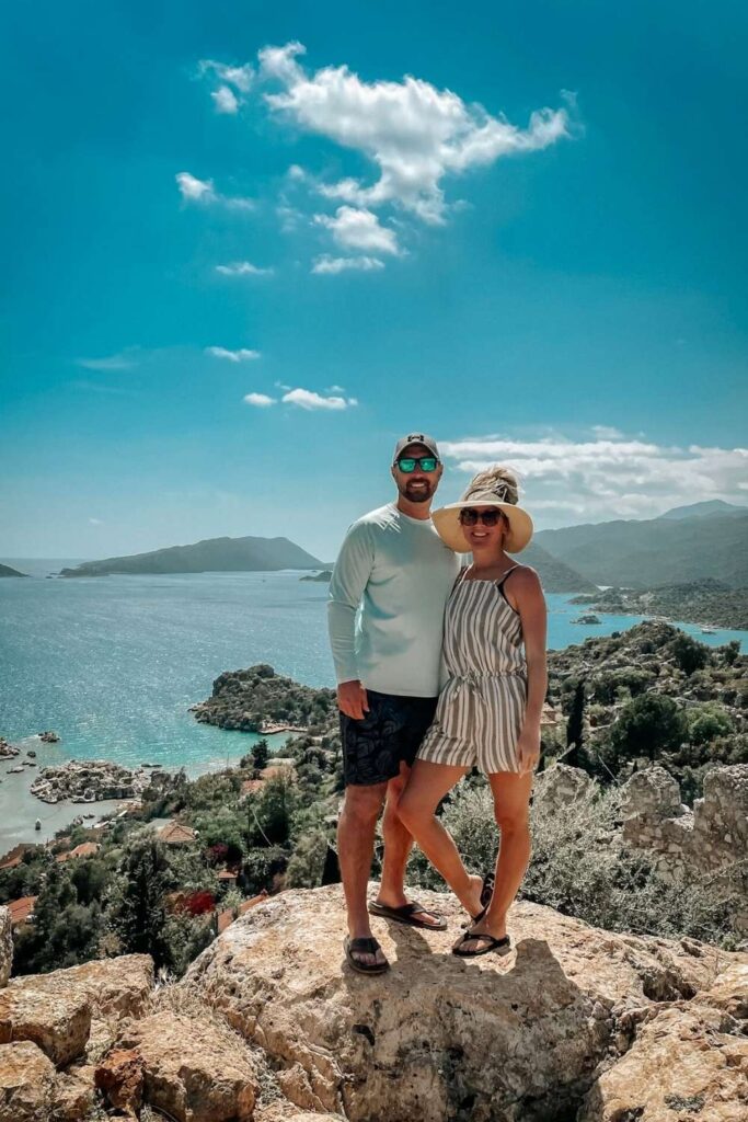 This photo showcases Kate and her husband enjoying the stunning views of the Turkish Riviera, standing on a rocky overlook with the turquoise sea and lush green hills in the background. The man wears casual beachwear and sunglasses, while Kate is dressed in a light summer outfit with a sun hat.