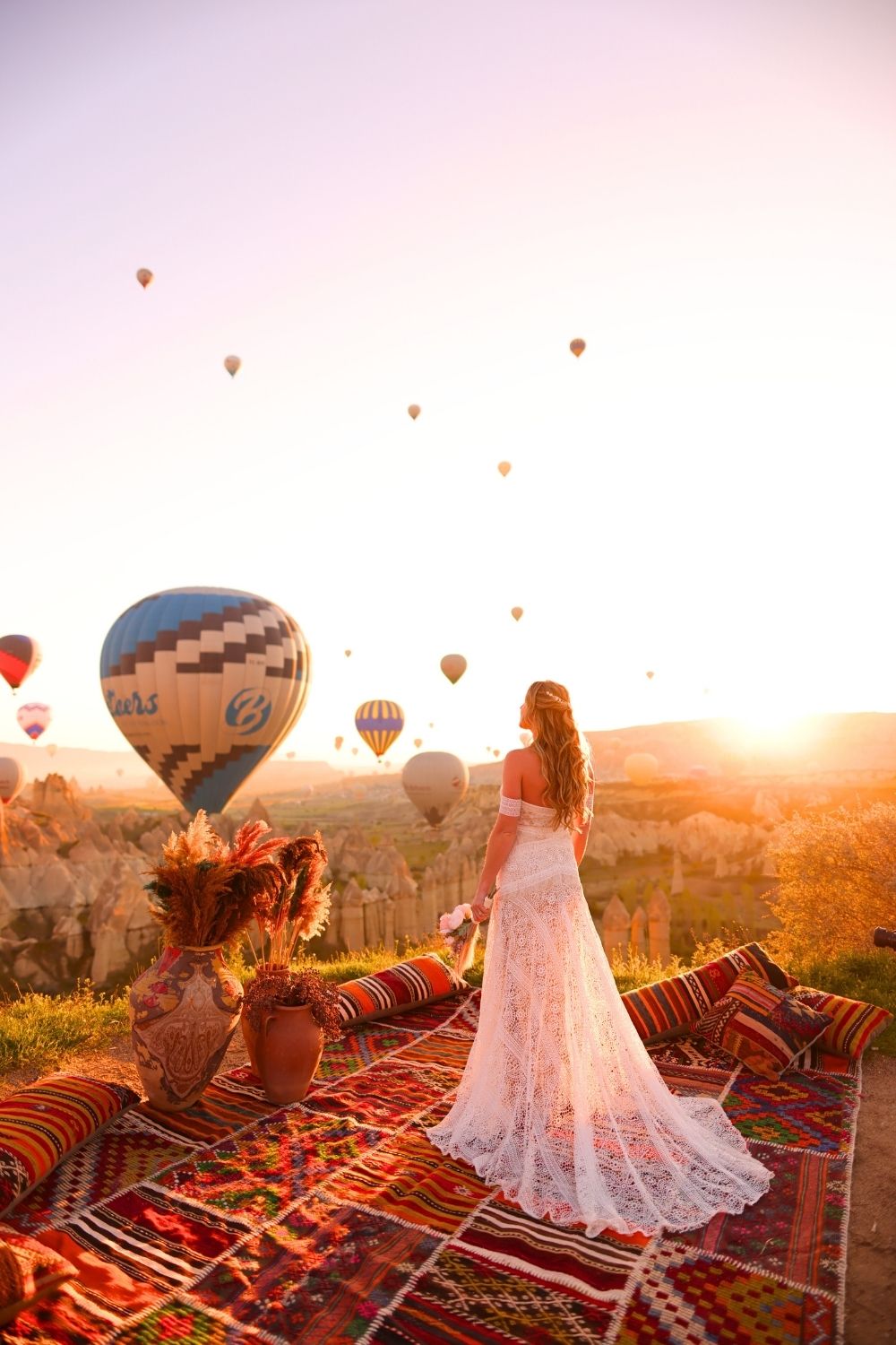 This image showcases a serene morning in Cappadocia, Turkey, with colorful hot air balloons floating above the unique rock formations. Kate in a flowing white dress, standing on a vibrant patterned rug adorned with vases and flowers, gazing at the sunrise and the breathtaking view.