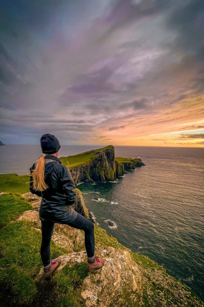 This photo captures the dramatic cliffs and the lighthouse at Neist Point on the Isle of Skye in Scotland during a vibrant sunset. Kate in black outdoor gear stands on a rocky outcrop, overlooking the sea and rugged landscape, with golden hues reflecting off the water. The scene showcases the natural beauty and serene atmosphere of this iconic Scottish location.