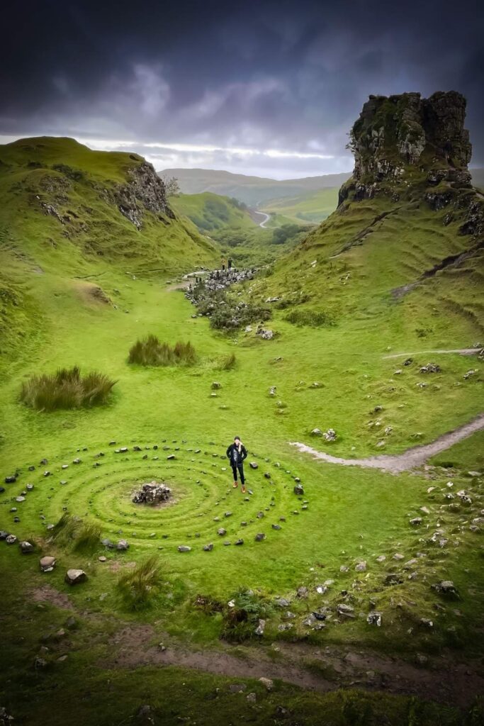 This image shows a lush green landscape in Scotland, featuring a spiral of stones in the foreground and dramatic rock formations in the background. A single person stands within the spiral, emphasizing the mystical and serene atmosphere of the location, under a moody, overcast sky.