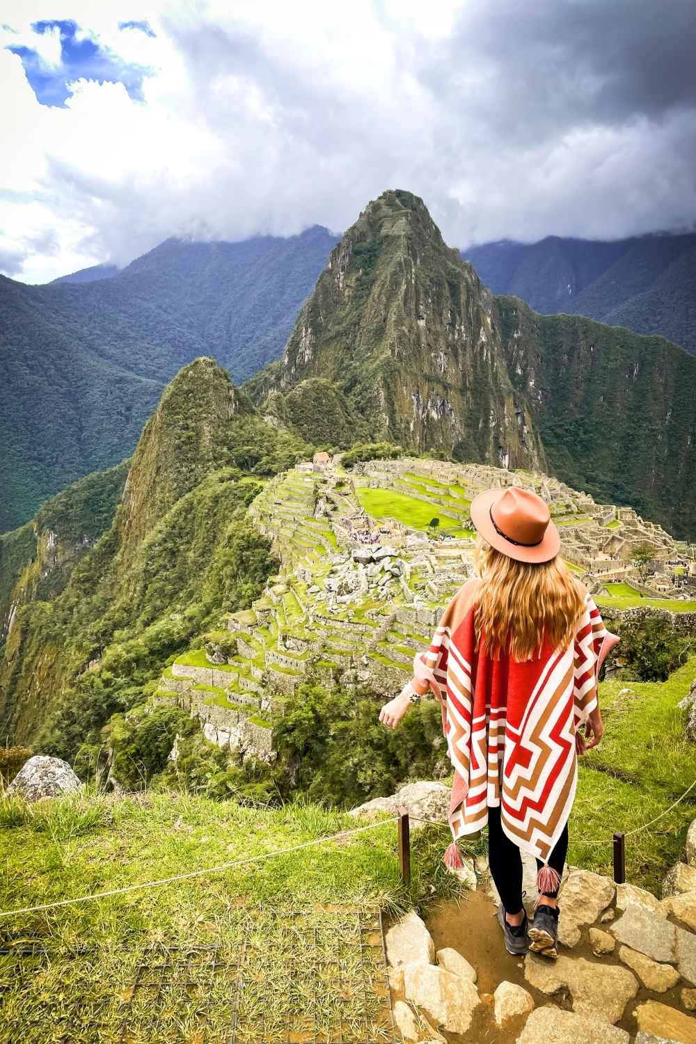 This image showcases the iconic Machu Picchu in Peru, with its ancient Inca terraces and structures nestled among lush green mountains. Kate is wearing a patterned red and white poncho and a brown hat stands on a stone path overlooking the archaeological site.