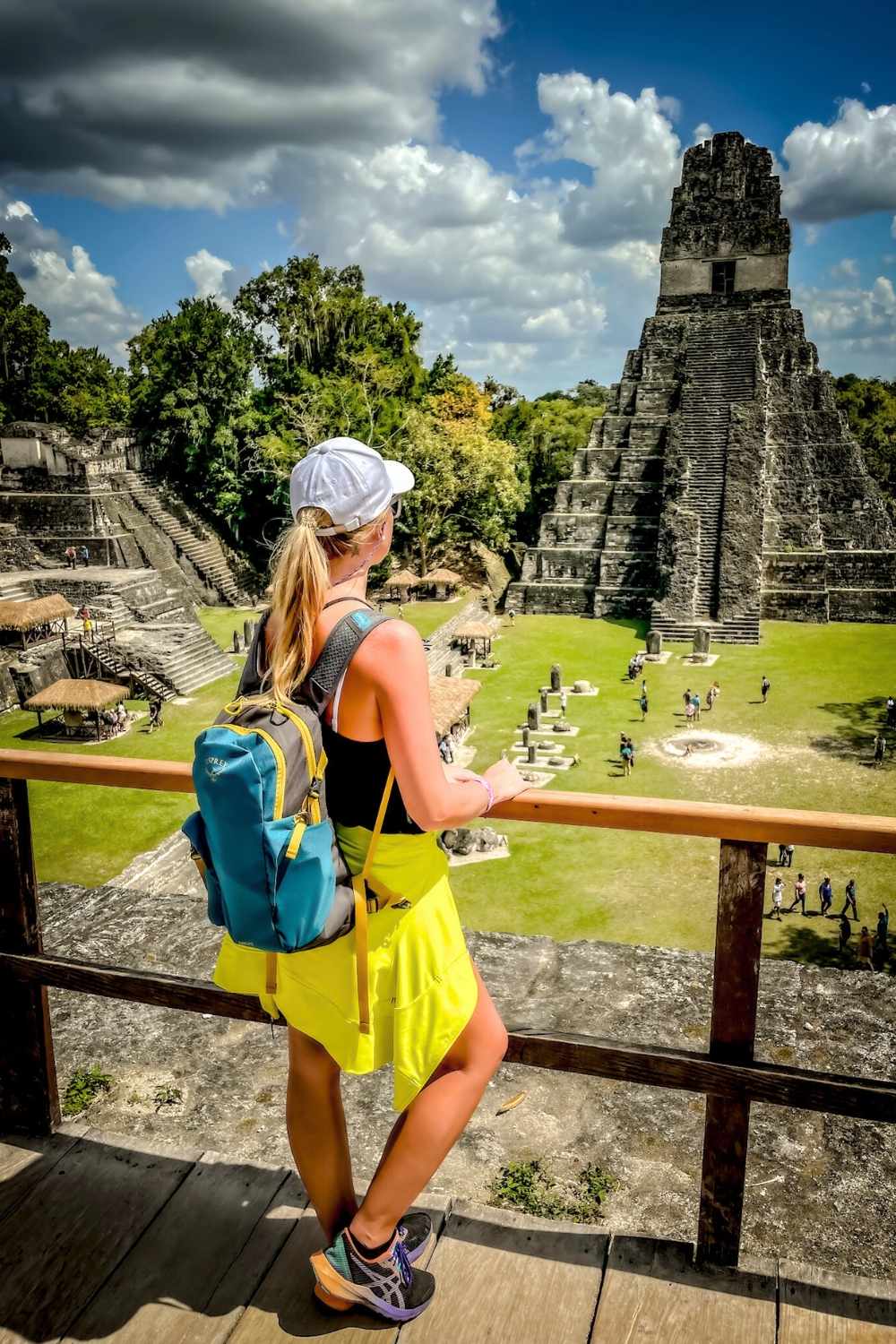 This image shows Kate overlooking the ancient Mayan ruins of Tikal, Guatemala, featuring a grand pyramid surrounded by lush jungle. Kate is wearing a white cap and backpack, gazing at the site’s iconic architecture and stone structures under a partly cloudy blue sky.