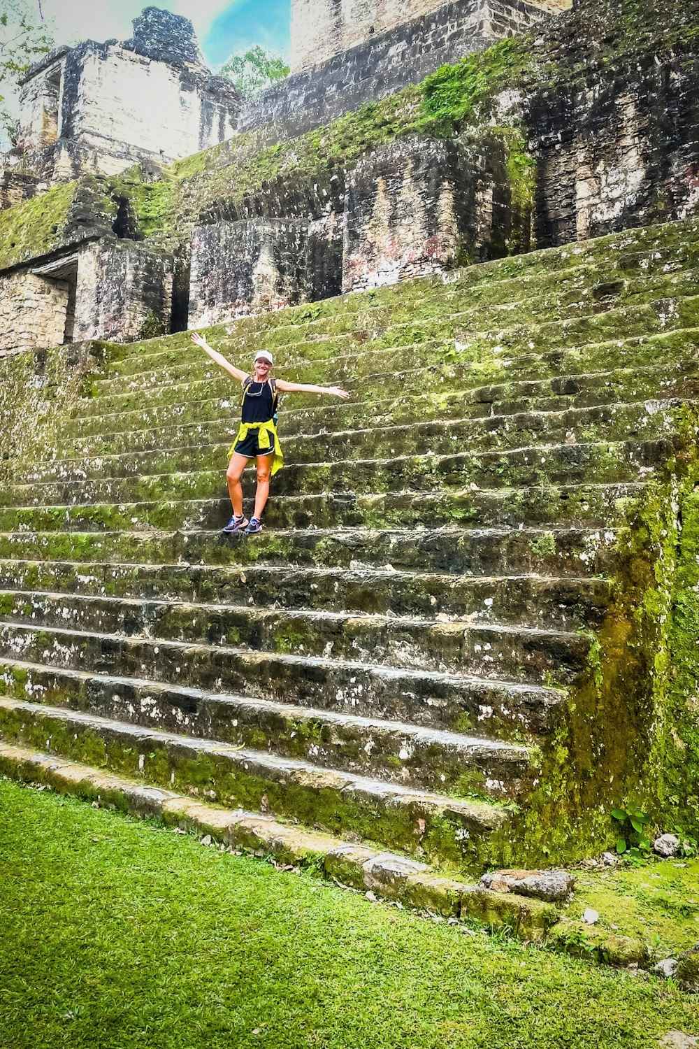 This image captures Kate joyfully posing on the moss-covered stone steps of an ancient pyramid at the Tikal archaeological site in Guatemala. The weathered structure, surrounded by lush greenery, showcases the grandeur of Mayan architecture.