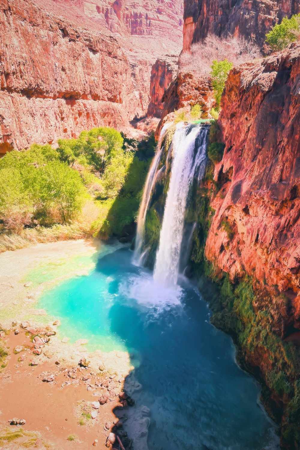 This image captures the stunning Havasu Falls, a vibrant turquoise waterfall cascading down red rock cliffs in the Grand Canyon, Arizona. Surrounded by lush green trees, the natural beauty of this iconic spot contrasts dramatically with the arid desert landscape, creating a serene oasis.