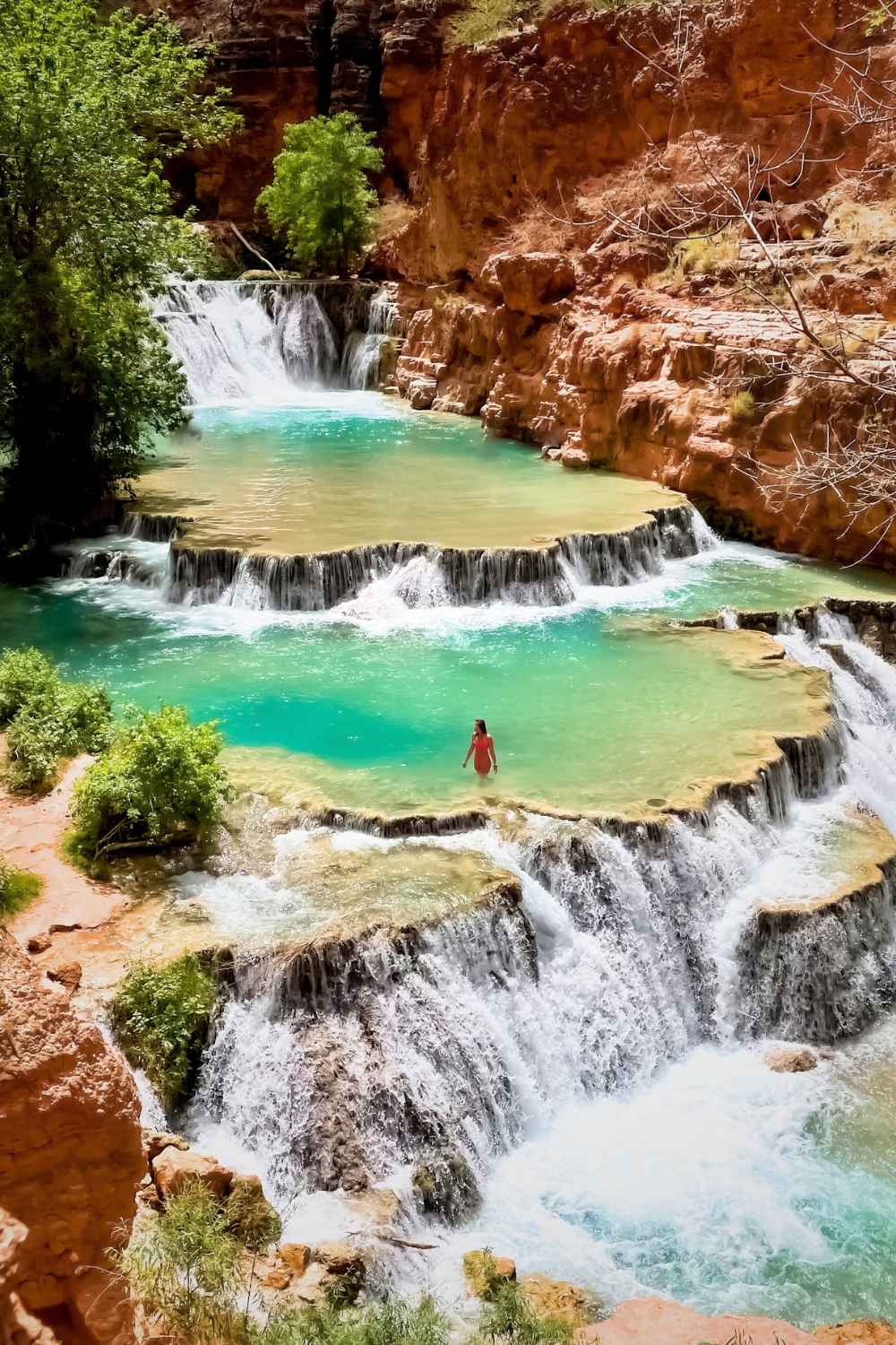 This image showcases Beaver Falls in Havasupai, Arizona, a series of cascading turquoise pools and waterfalls surrounded by vibrant red rock cliffs. A lone person in a red swimsuit wades in one of the serene pools, highlighting the natural beauty and tranquility of this iconic desert oasis.