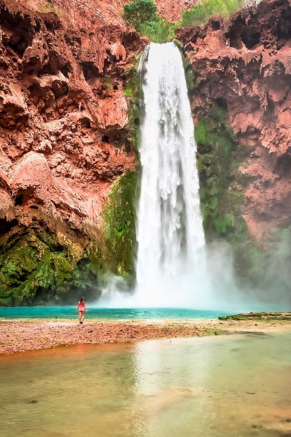 This image showcases Mooney Falls in Havasupai, Arizona, a dramatic waterfall plunging down red rock cliffs into a turquoise pool surrounded by lush greenery. A lone person in a pink outfit stands near the water, emphasizing the immense scale and beauty of the cascading falls.