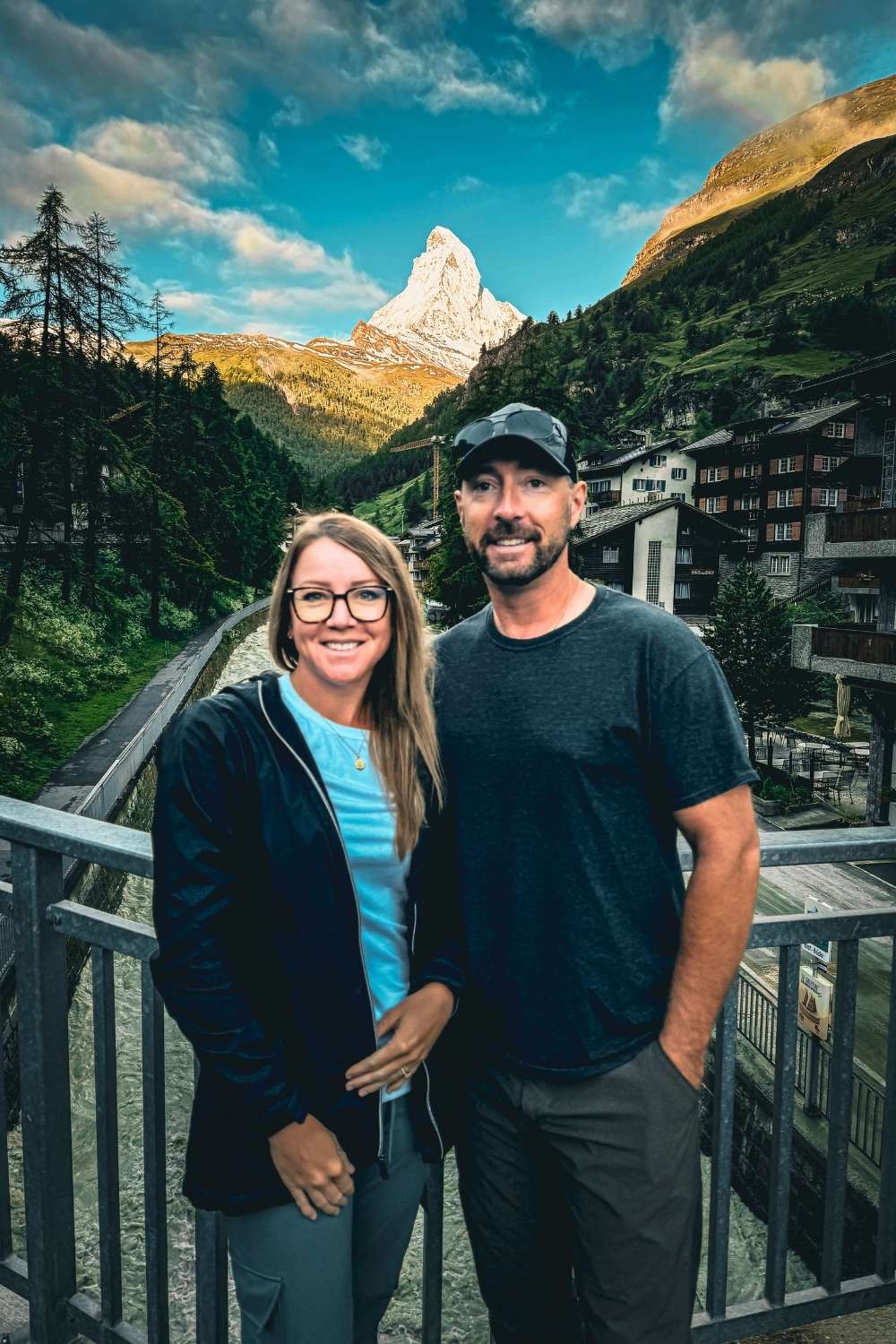 This photo features Kate and her husband smiling on a bridge in Zermatt, Switzerland, with the iconic Matterhorn peak illuminated in the background. The picturesque scene includes traditional alpine chalets and a flowing river framed by lush green hills and the dramatic mountain landscape.