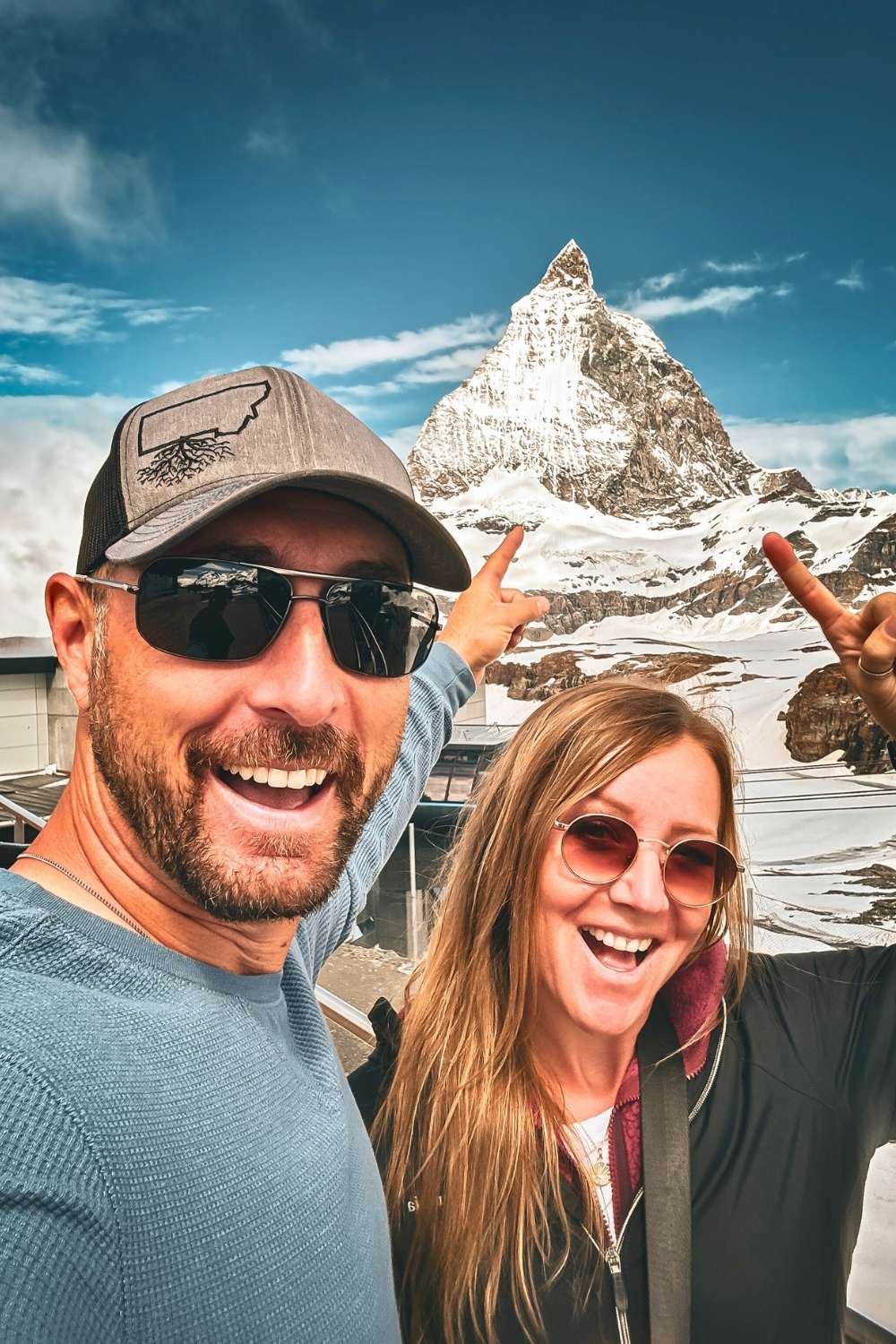 This photo shows Kate and her husband smiling excitedly and pointing at the iconic Matterhorn in Zermatt, Switzerland. The snow-covered peak stands majestically under a clear blue sky, framed by their enthusiastic expressions and gestures.