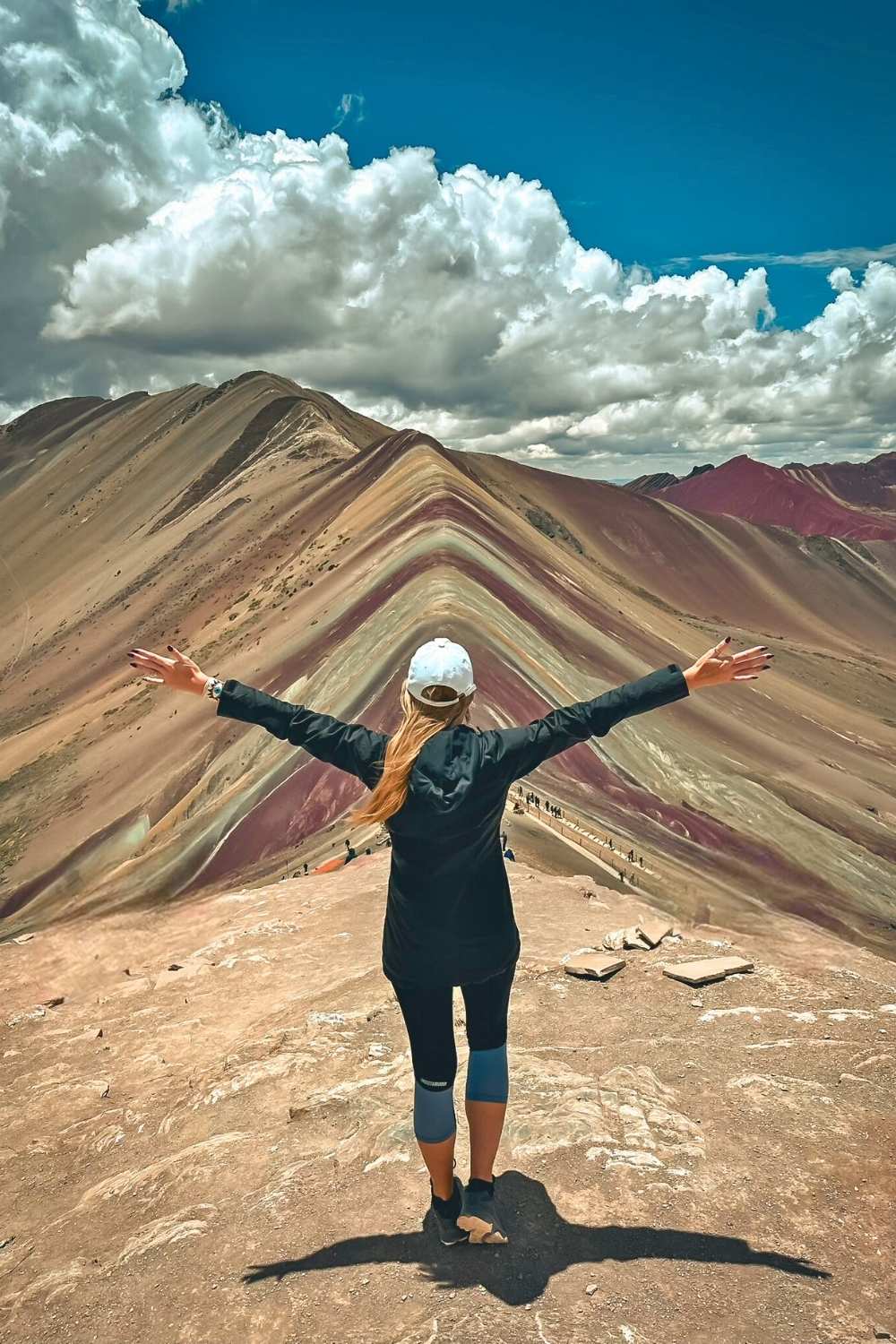 This image features Kate standing with arms outstretched, overlooking the vibrant, layered hues of Rainbow Mountain (Vinicunca) in Peru. The colorful mountain ridge contrasts beautifully with the dramatic clouds and blue sky above, capturing the natural splendor of this iconic Andean landscape.